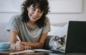 Glad woman with cat writing in planner while using laptop