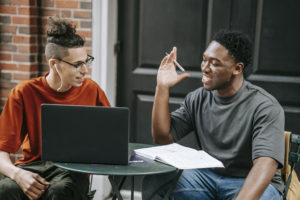 Positive diverse male students browsing laptop while studying together in street cafe